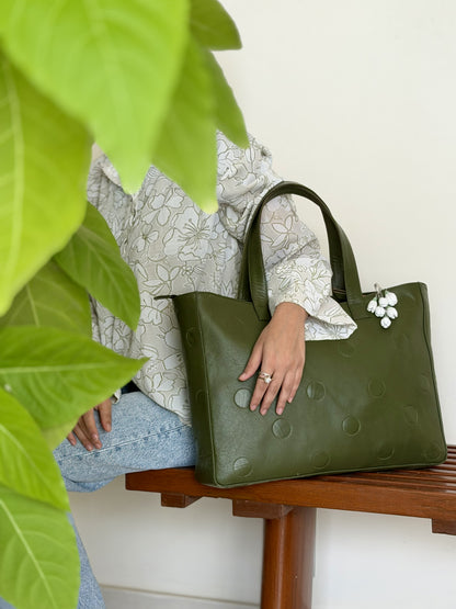 An olive green textured leather tote bag with circular debossed details, held by a person sitting on a wooden bench. The person is wearing a white floral long-sleeve top and light blue jeans, with a green houseplant in the foreground and a white wall in the background.