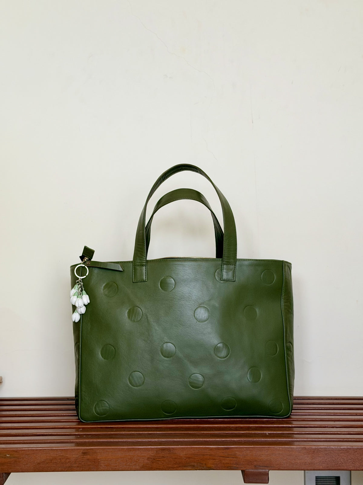 An olive green leather tote bag with debossed circular dot patterns and dual top handles, resting on a wooden bench against a plain off-white wall.