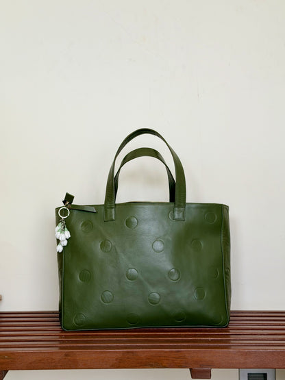 An olive green leather tote bag with debossed circular dot patterns and dual top handles, resting on a wooden bench against a plain off-white wall.