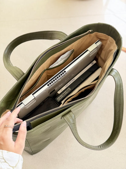 An olive green leather tote bag shown open, filled with a silver laptop, notebook, and other accessories. A hand is visible holding the bag open, set against a neutral beige background.