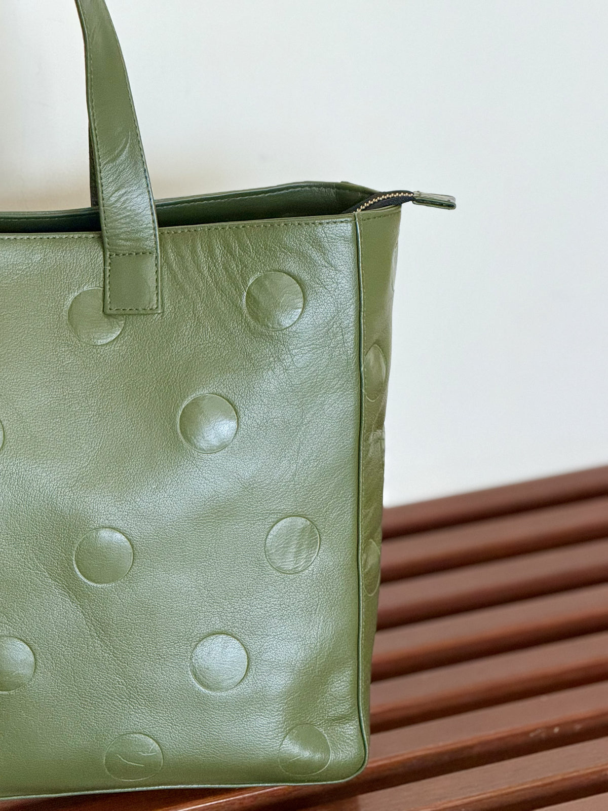 An olive green leather tote bag with a unique debossed circular dot pattern, resting on a brown wooden slatted bench against a plain off-white wall.