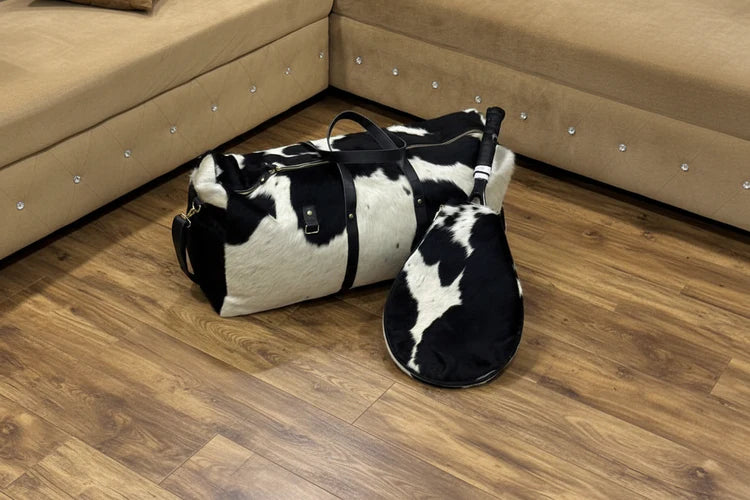 black and wite cowhdie leather tennis duffel bag with a tennis recquit , on an oak wood wooden floor in a room