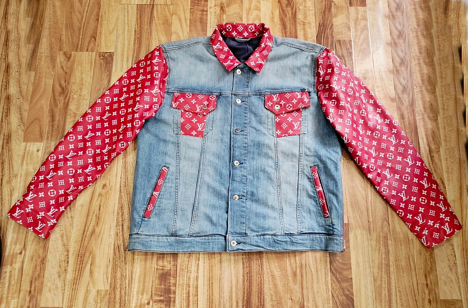 trucker Denim jacket with white designer monogram pattern on red leather sleeves and pockets laying flat on a wooden surface